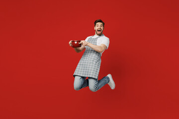 Full body young happy fun surprised male housewife housekeeper chef cook baker man wear grey apron holding in hand saucepan looking camera isolated on plain red background studio Cooking food concept