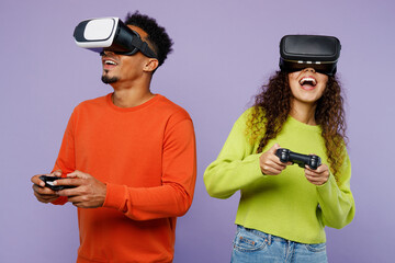 Young couple two friend family man woman of African American ethnicity in casual clothes together watch in vr headset pc gadget play pc game with joystick console isolated on plain purple background