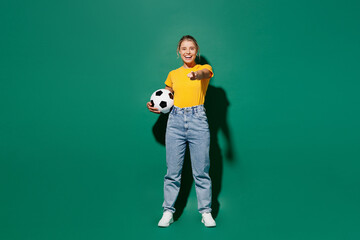 Naklejka na ściany i meble Full body young woman fan wear yellow t-shirt cheer up support football sport team hold in hand soccer ball watch tv live stream point finger camera on you encourage isolated on dark green background.