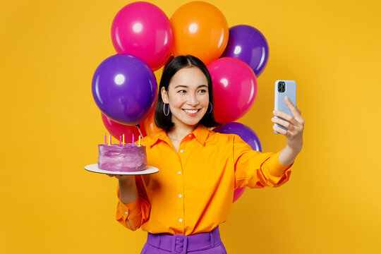 Happy fun young woman wear casual clothes celebrate near balloons hold cake with candles do selfie shot on mobile cell phone isolated on plain yellow background. Birthday 8 14 holiday party concept