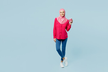 Full body young arabian asian muslim woman wear pink abaya hijab hold takeaway delivery paper cup coffee to go isolated on plain pastel light blue cyan background People uae islam religious concept.