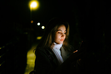 Young caucasian woman using mobile phone outdoor in the evening, walking on city streets.