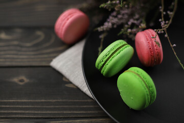 traditional American macaroons in pink and bright green color on a wooden background and a black plate next to purple fragrant flowers