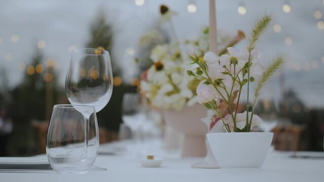 Close-up of festive wedding table setting with wine glass, pastel wild flowers and spikelets at small cozy wedding party, retro lamps on the background, slow motion.