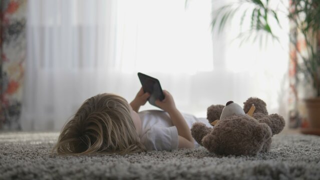 Little boy watching smartphone lying on carpet near teddy bear, floor view