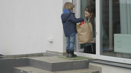 Funny little child give a paper bag with groceries to mother sitting at door,