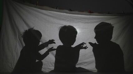 Three children playing shadows on sheet tent lit by lamp in their room