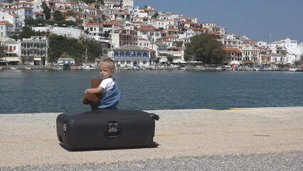 Child holding a teddy bear sitting on suitcase and admiring white sea town