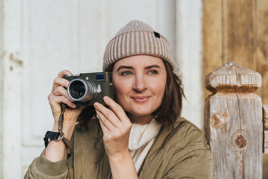 Happy smiling girl wearing beanie hat looking away while taking pictures by vintage photo camera. Portrait of young travel woman on vacation.