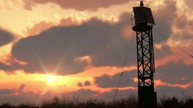 The openwork silhouette of the lighthouse against the backdrop of a cloudy sunset sky among tall grass, which is swayed by the wind. A beacon light in the wilderness at the edge of the world.