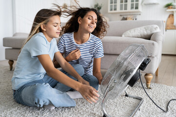 Mother and daughter cooling down near electric fan blowing fresh air, spending time together at...