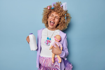 Emotional pregnant woman holds bottle with milk and doll screams loudly carries net bag on shoulder dressed in casual clothes prepares for maternity isolated over blue background. Pregnancy concept