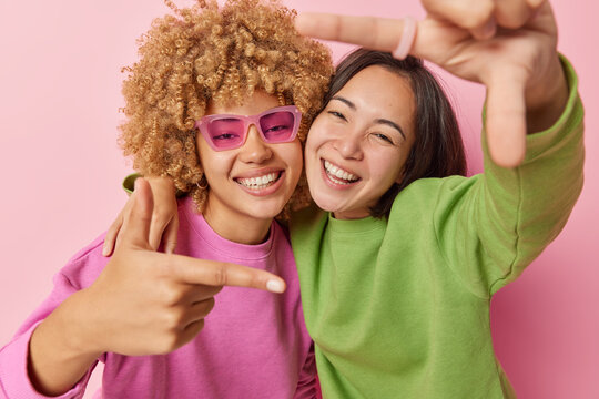 Best female friends have fun make frame gesture with fingers smile toothily wear casual jumpers foolish around isolated over pink background. Happy biracial women measure angle picture something - Powered by Adobe