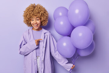 Festive occasion concept. Positive curly haired woman wears raincoat points at bunch of inflated balloons celebrates bithday isolated over purple background. Female model enjoys celebration.