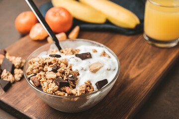 Granola cereal oatmeal with white yogurt, chocolate, banana fruit and nuts in a bowl on dark wooden board