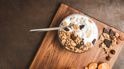 Granola cereal oatmeal with white yogurt, chocolate, fruist and nuts in a bowl on dark wooden board, top view