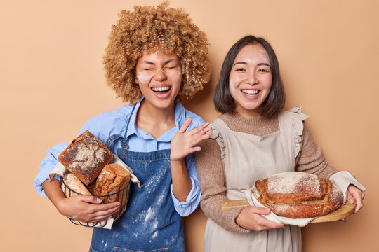 Positive two women stand happy with fresh delicious bread smeared with flour wear aprons satisfied after finishing baking isolated over brown background. Female bakers work at bakery factory