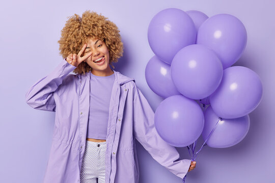 Horizontal shot of positive woman with curly hair makes peace sign holds inflated balloons gathered in bunch dressed in outerwear poses against purple background celebrates special occasion.