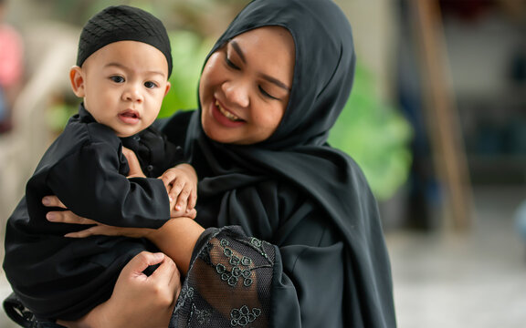 Portrait Adult muslim mother wearing traditional black dress and headscarf, carrying little baby boy, smiling with happiness in indoor home with copy space. Education, Family, Religious Concept.