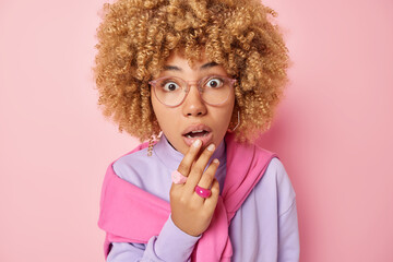 Image of shocked curly haired woman keeps hand on lips keeps mouth opened stares speechless at camera realizes something amazing wears transparent eyeglasses and pullover isolated over pink background