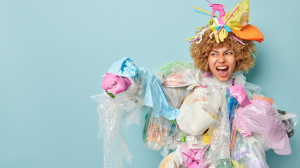 Emotional annoyed curly haired woman clenches fist and screams loudly about pollution on Earth covered with plastic garbage ready to save planet from contamination isolated over blue background
