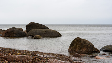 Fototapeta na wymiar In the seascape, glacial granite stones and boulders are washed by the calm waters of the Baltic Sea. Inclement cloudy weather, drizzle. Cloud haze gently covers the horizon