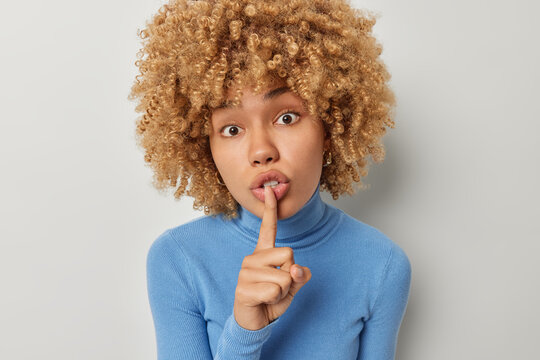 Portrait of surprised curly haired woman hides secret presses figer to lips shushes at camera wears casual blue turtleneck isolated over grey background asks keep voice down shares interesting rumor