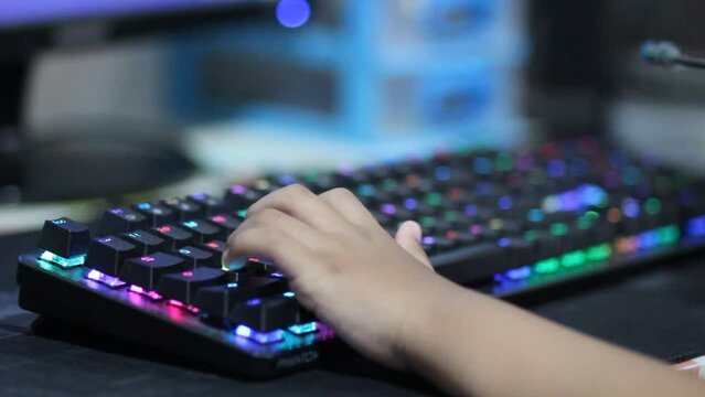 Closeup video of a child's hands pressing keys on a computer keyboard playing an online game with deliberately blurred background and colorful LED lights on the keyboard.