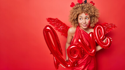 Horizontal shot of pleased curly haired woman holds inflated letters meaning love wears elegant dress concentrated somewhere isolated over red background blank space for your promotional content
