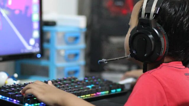 A little boy plays online games on a computer using a headset and a mechanical keyboard with colorful led lights.