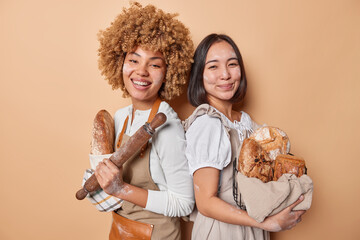 Happy women baked delicious crusty bread holds rolling pin stnad backs smile broadly sell homemade breadwork at bakery store smeared with flour isolated over brown background. Domestic production