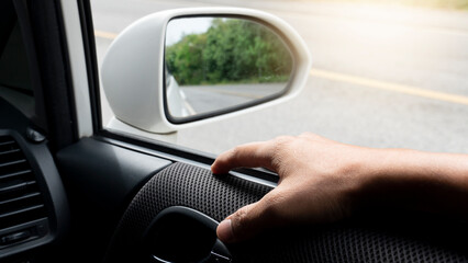 Driver's hands rest on the top of the car door. Mirror wing of white car with reflection of asphalt road and green forest.