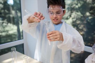 Selective focus on the hands of a smart schoolboy in safety goggles and white lab gown, pouring reagent into a test tube, conducting scientific chemical experiment in a school chemistry laboratory