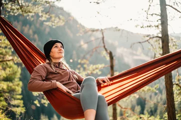 Fotobehang Woman with cap resting in comfortable hammock during sunset. Relaxing on orange hammock between two trees pine enjoying the view © alexanderuhrin