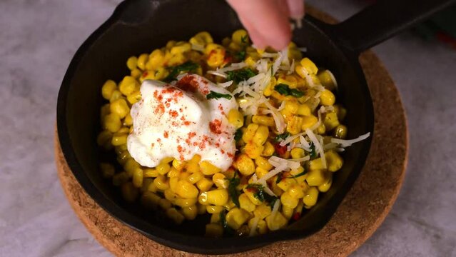 Esquites, grilled corn, sprinkled with grated cheese and served with sour cream and chili powder in cast iron pan. Mexican street food meal