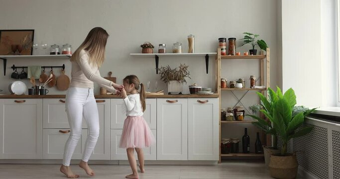 Happy young mother hold hands dancing with preschooler adorable daughter in festive fluffy pink skirt, moving to music barefoot in modern warm domestic kitchen, having fun on weekend celebrate event