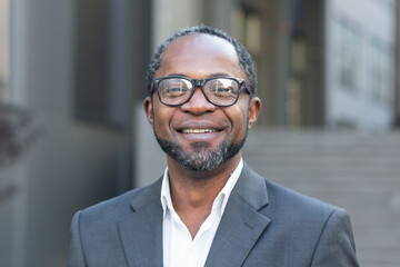 Close-up photo. Portrait of a successful handsome African American man. A businessman, lawyer, boss stands in front of the office center in a suit and glasses, looks at the camera, smiles.