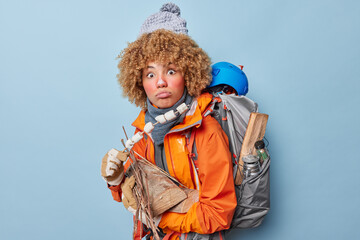 Photo of surprised adult woman camper carries wood and roasted marshmallow wears knitted hat orange jacket spends free time actively poses with rucksack on back isolated over blue background