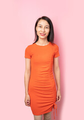 Fototapeta premium Asian brunet woman in orange dress on pink background looking at camera. 