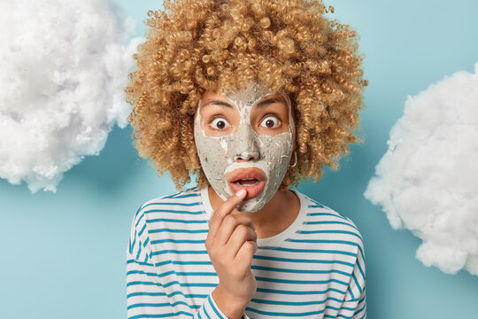 Portrait of surprised woman with curly hair keeps finger on lips applies facial clay mask for skin treatment wears casual striped jumper isolated over blue background white clouds above. Face care