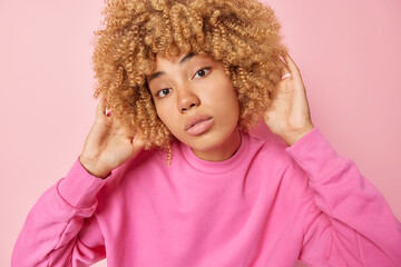 Headshot of beautiful woman with curly bushy hair keeps hands on head looks directly at camera has calm expression clean healthy skin wears pullover isolated on pink background. Natural beauty concept
