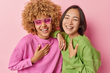 Overjoyed multiracial women laugh happily smile toothily keep hands on chest have upbeat mood wear sunglasses casual jumpers stand closely to each other isolated on pink background. Positive emotions