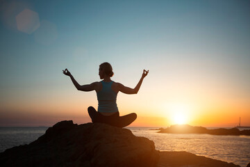 A woman does yoga in the lotus pose, meditating on the coast during sunset.