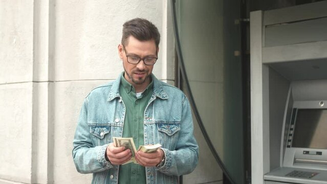 Portrait of happy rich young adult businessman counting dollar notes withdrawn from bank card using ATM terminal outside. Beautiful cheerful male holding money smiling. Finance concept.