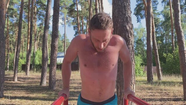 A young athletic man with a naked torso, dressed in blue shorts, does push-ups on the uneven bars at a training ground in the forest. The concept of a zor lifestyle. Summer
