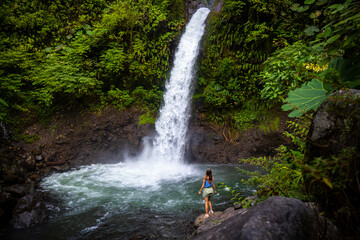 A beautiful girl stands on rocks under a powerful tropical waterfall in Costa Rica; la paz...