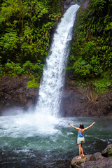 A beautiful girl stands on rocks under a powerful tropical waterfall in Costa Rica; la paz waterfall in the jungle