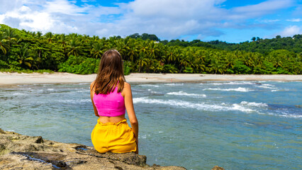 A beautiful girl in colorful clothing sits on the rocks admiring a tropical beach with palm trees;...