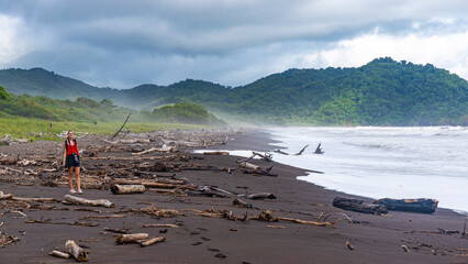 girl walking along Costa Rica's black sand beach during a storm; large tree trunks tossed onto the beach by a hurricane