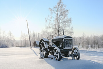 An old tractor stands in deep snowdrifts, covered with snow.Village in winter on a sunny frosty day. © Lexis_Jan